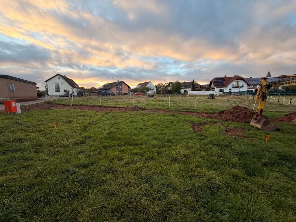 Blick über die Wiese mit den orangen Pflöcken der Grobabsteckung und einem frisch abgeschobenen Streifen, rechts die Baggerschaufel, im Hintergrund die Nachbarhäuser