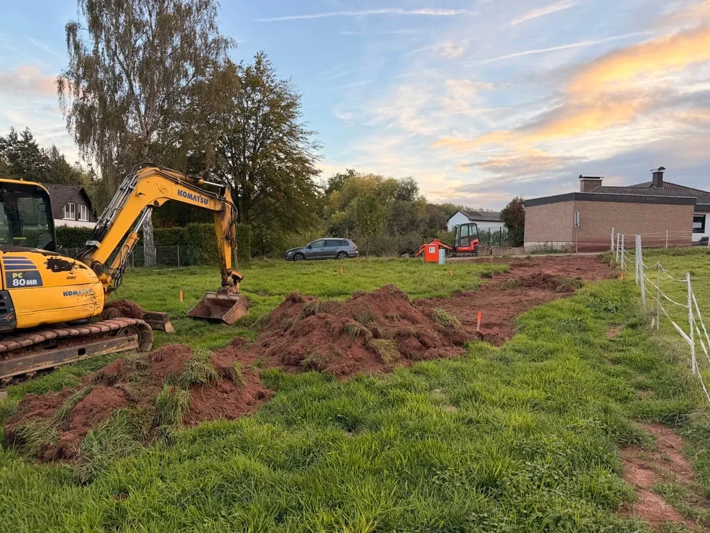 Ein gelber Komatsu-Bagger auf unserem Grundstück, davor aufgeschobene Erdhaufen mit Grasnarbe, rechts der Weidezaun, darüber ein Abendhimmel