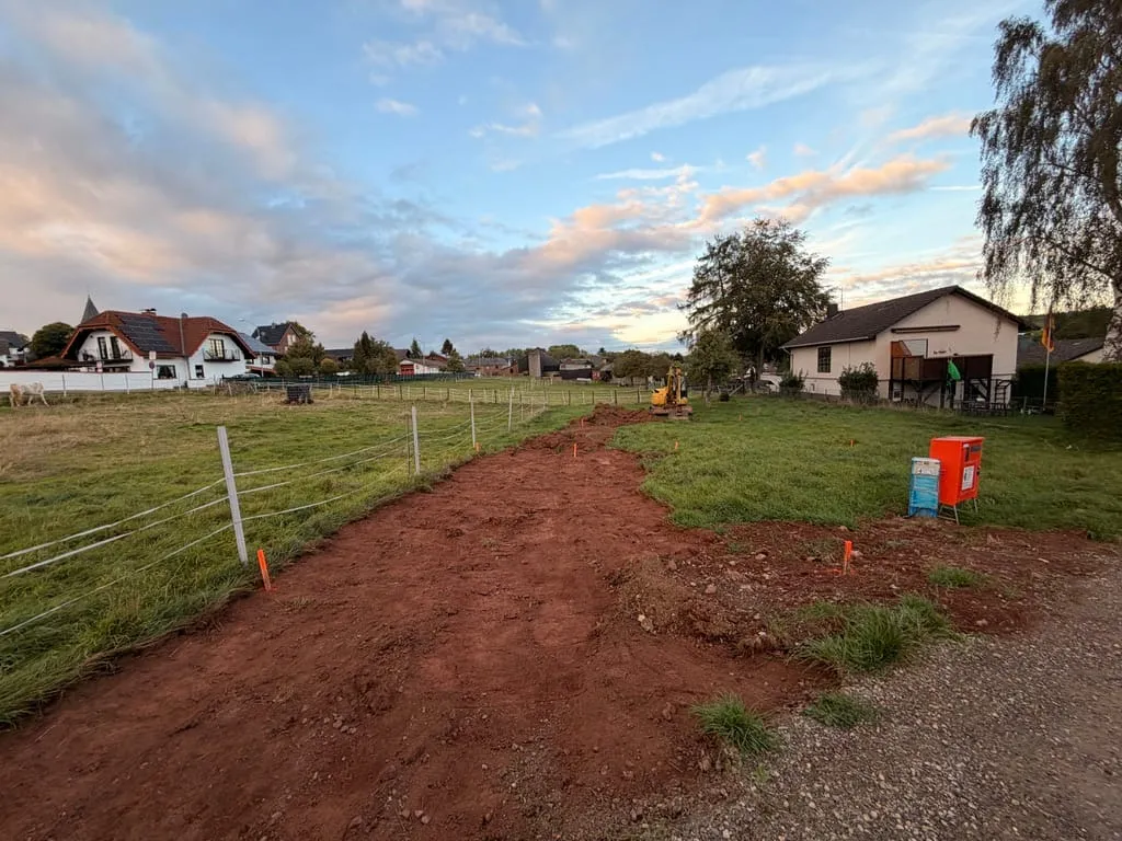 Ein breiter, frisch abgeschobener Streifen roter Erde führt vom Schotterweg über das Grundstück, rechts der orange Baustromkasten, hinten der Bagger