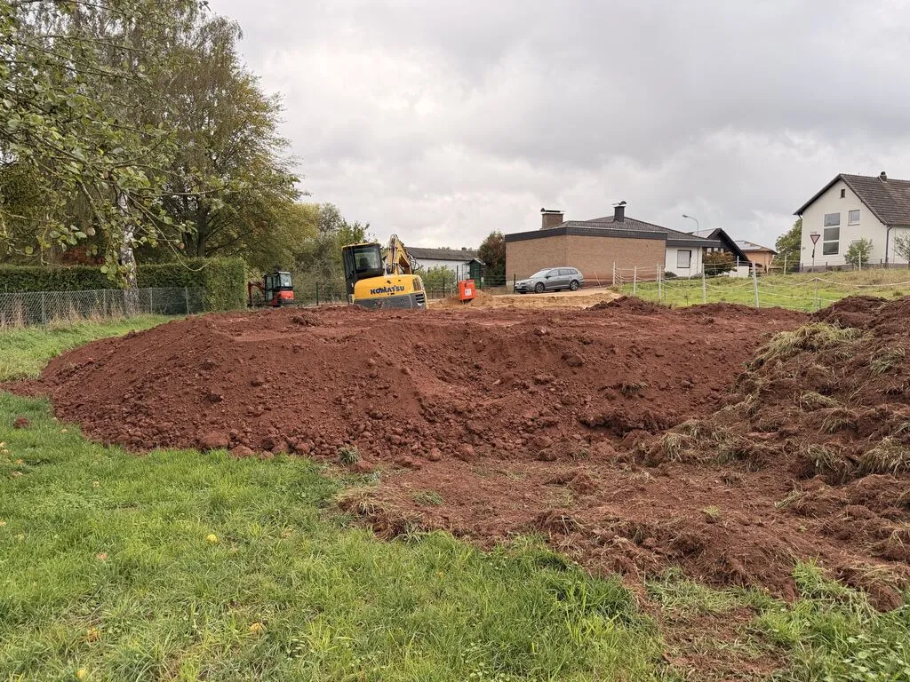 Ein langer Haufen roter Erde mit Grasbüscheln, dahinter der Komatsu-Bagger und der Baustromkasten