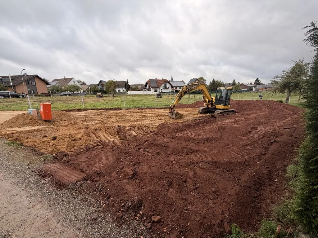 Blick vom Schotterweg: links der orange Baustromkasten und der helle Kies, rechts eine breite Fläche roter Erde mit Kettenspuren, dahinter der Bagger und die Häuser im Dorf